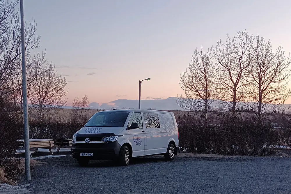 Ein weißer Campervan, der bei Sonnenuntergang auf dem Campingplatz Búðardalur in Island geparkt ist, mit einer Bank und einer Laterne in der Nähe, erinnert an die heitere Stille des Wintercampings.