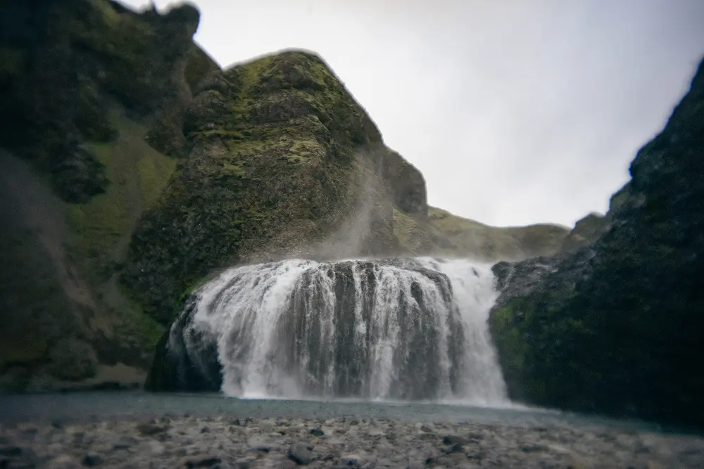 Ein breiter Wasserfall stürzt in Kaskaden über dunkle Felsen, die von moosbewachsenen Klippen umgeben sind, und bei bedecktem Himmel steigt Nebel aus dem fallenden Wasser auf.