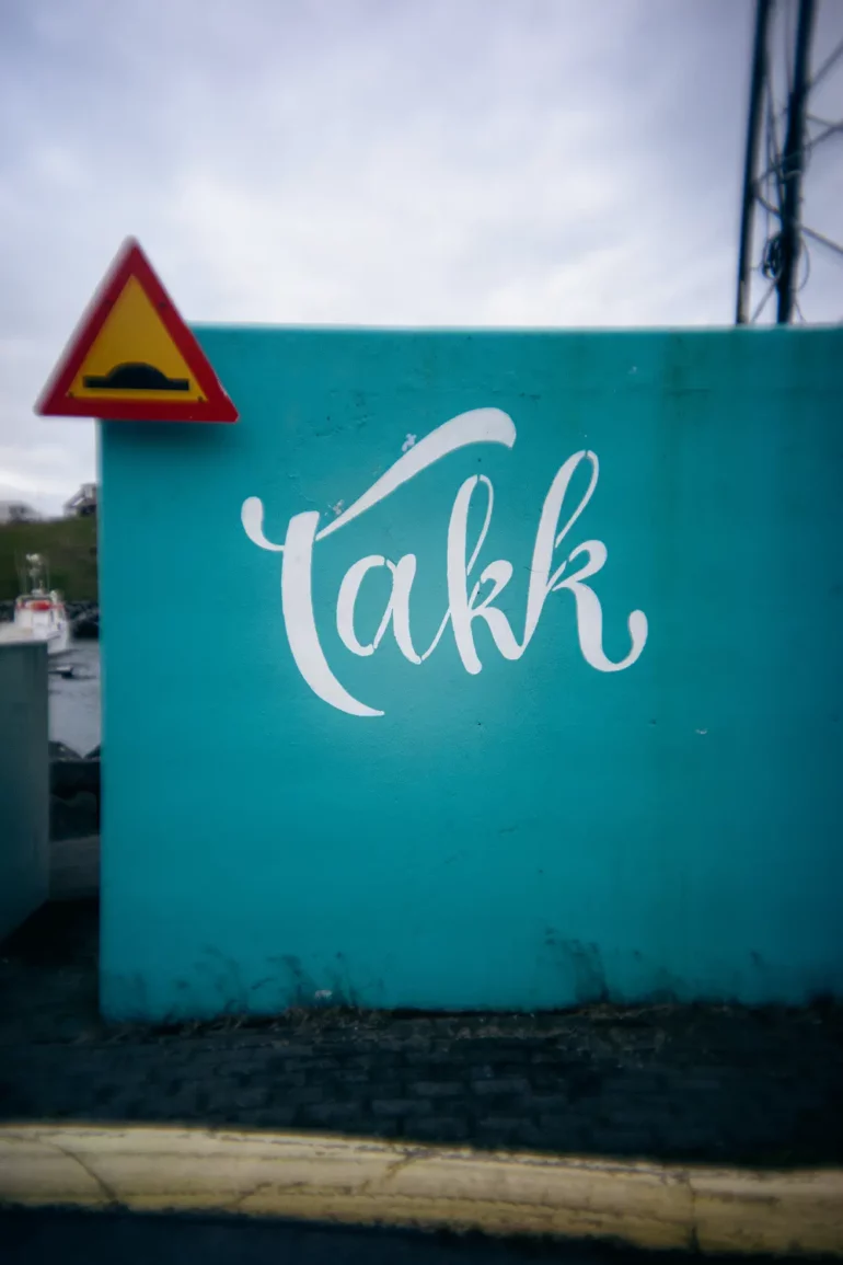 Eine blaue Wand mit dem in weißer Schrift gemalten Wort Takk. Oberhalb der Mauer befindet sich ein Straßenschild mit einer Bodenwelle. Der Boden ist mit Pflastersteinen gepflastert und am unteren Ende ist ein Bordstein zu sehen.