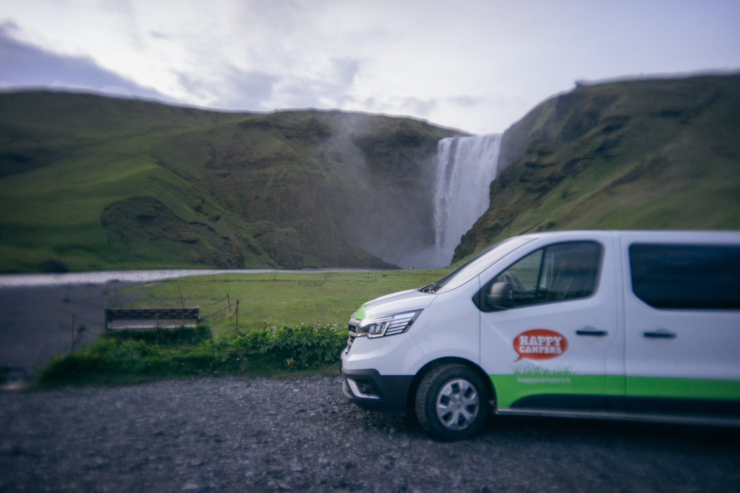 Ein weißer Lieferwagen mit Happy-Camper-Schriftzug parkt auf einer Kiesfläche vor einer Graslandschaft und einem großen Wasserfall und fängt die Essenz des Campings in Island unter einem bewölkten Himmel ein.