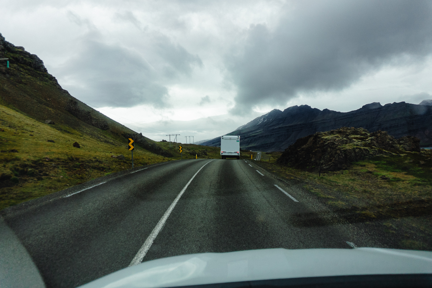 Eine zweispurige Straße schlängelt sich unter wolkenverhangenem Himmel durch eine bergige Landschaft. Vor uns fährt ein weißes Fahrzeug - vielleicht auf dem Weg zum Camping auf einer Insel - und auf der linken Seite sind gelbe Straßenschilder zu sehen.