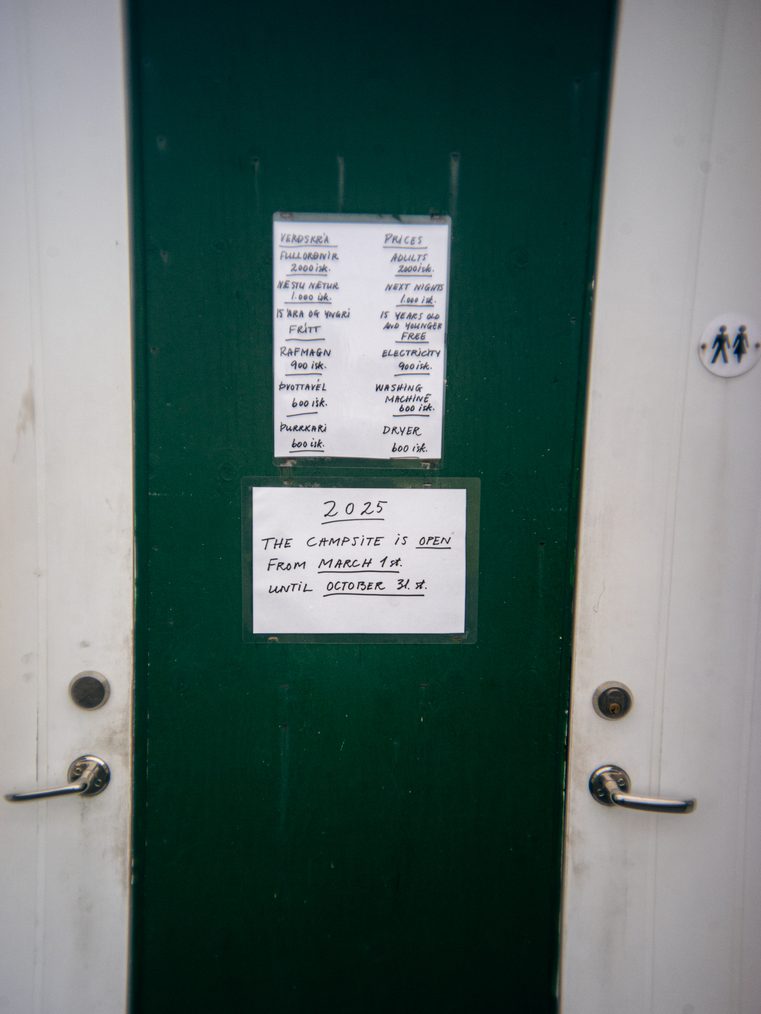 Eine grüne Tür mit zwei weißen Schildern heißt die Campinggäste auf der Insel willkommen. Auf dem oberen Schild sind Namen und Zuweisungen aufgeführt, während das untere Schild darauf hinweist, dass der Campingplatz vom 1. März bis zum 31. Oktober geöffnet ist. Auf der rechten Seite befinden sich zwei silberne Griffe und ein Toilettensymbol.
