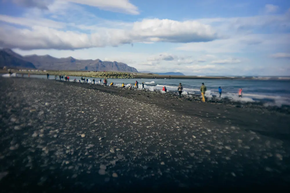 Ein felsiger Strand mit vereinzelten Menschen, die am Ufer spazieren gehen. Der Himmel ist größtenteils bewölkt, und im Hintergrund sind die Berge auf der anderen Seite des Wassers zu sehen.