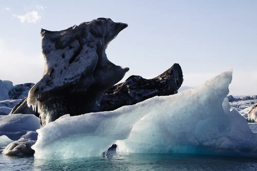 Große, unregelmäßig geformte Eisberge treiben im ruhigen Wasser, wobei ein Eisberg einen Bogen bildet und ein anderer zerklüftet dahinter aufragt, vor dem Hintergrund eines blassen Himmels und einer fernen Schneelandschaft.