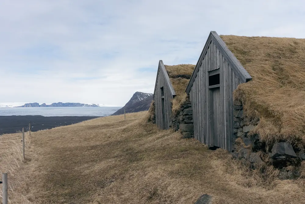 Zwei kleine Holzhäuser mit grasbedeckten Dächern sind in einen grasbewachsenen Hang gebaut, von dem aus man in der Ferne schneebedeckte Berge und einen Gletscher unter einem bewölkten Himmel sehen kann.
