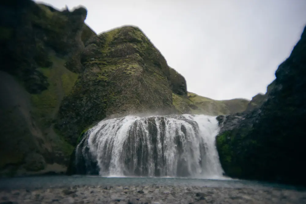 Ein breiter Wasserfall fließt über einen Felsvorsprung in ein ruhiges Becken, umgeben von moosbewachsenen Hügeln unter einem bedeckten Himmel.