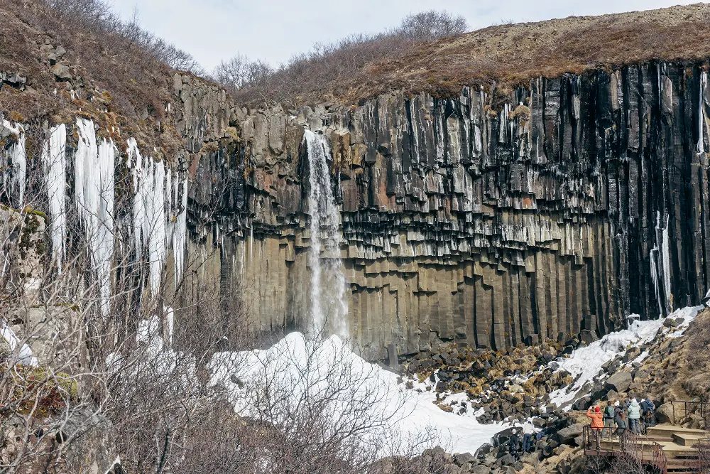 Ein hoher Wasserfall stürzt eine Klippe aus dunklen Basaltsäulen mit Eiszapfen und Schnee am Fuße hinunter. Laublose Sträucher und eine Gruppe von Menschen stehen in der Nähe einer Aussichtsplattform im Vordergrund.
