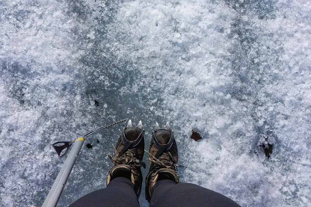 Eine Person, die Steigeisen trägt, steht auf einer vereisten, schneebedeckten Fläche und hält einen Trekkingstock. Das Bild ist aus dem Blickwinkel der Person aufgenommen, die auf ihre Füße und das umgebende Eis hinunterblickt.