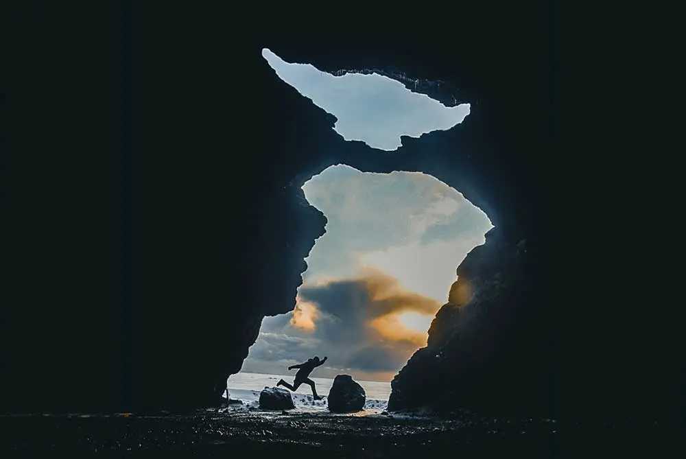 Eine Person ist als Silhouette zu erkennen, während sie zwischen Felsen in einer großen Höhle springt. Die Höhlenöffnung gibt den Blick frei auf einen bewölkten Himmel und schwaches Licht, das entweder vom Sonnenaufgang oder vom Sonnenuntergang stammt. Die Höhlenwände rahmen die Szene ein.