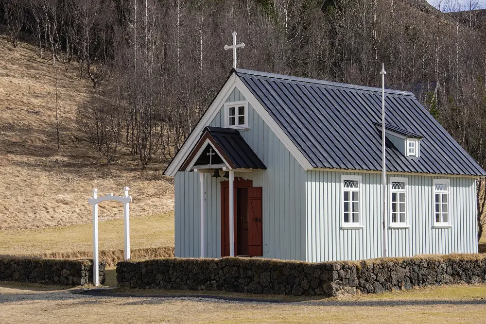 Eine kleine, hellblaue Holzkirche mit dunklem Dach und weißer Umrandung steht hinter einer niedrigen Steinmauer auf einer grasbewachsenen Wiese, mit kahlen Bäumen und einem Berghang im Hintergrund.