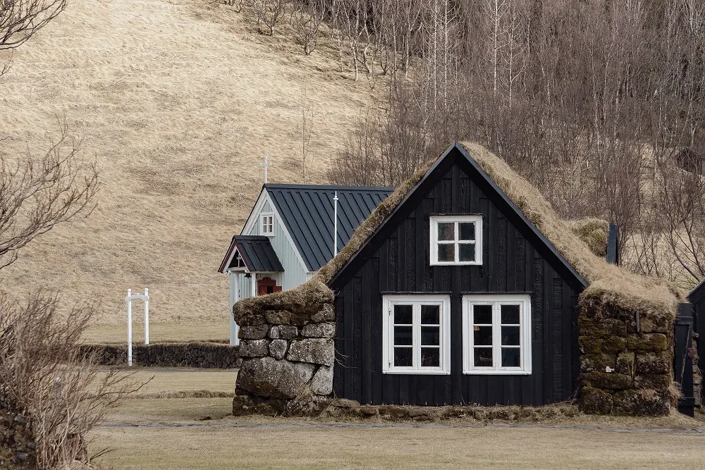 Ein kleines schwarzes Holzhaus mit weißen Fenstern und einem grasbedeckten Dach steht vor einem anderen ähnlichen Haus in einer grasbewachsenen, spärlich bewachsenen Landschaft mit trockenen Hügeln und kahlen Bäumen im Hintergrund.