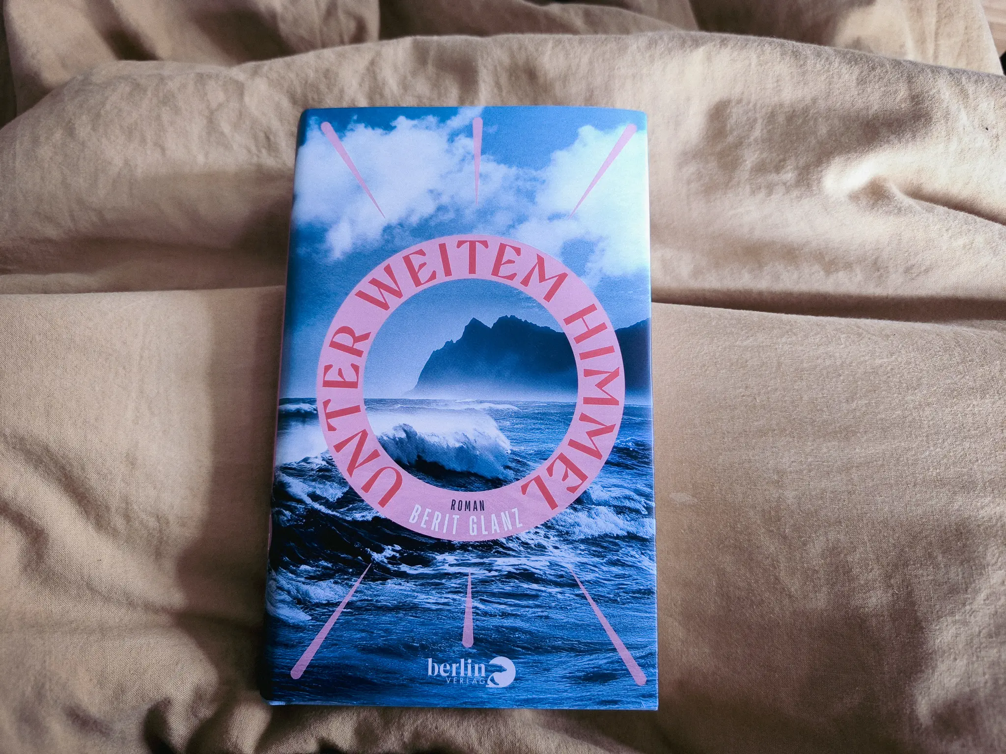 Ein Buch mit dem Titel Unter Weitem Himmel von Berit Glanz liegt auf einer beigen Stoffoberfläche. Der Einband zeigt eine Welle, Berge und Wolken mit dem Titel in rosa Buchstaben, die in einem kreisförmigen Muster angeordnet sind.