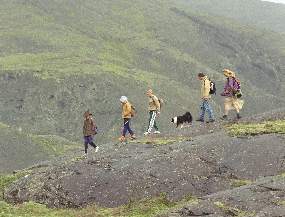 Fünf Menschen und ein Hund wandern im Gänsemarsch über einen felsigen Pfad an einem grünen Berghang, ihr Weg verkörpert die Liebe, die bleibt. Einige tragen Rucksäcke, während sie gemeinsam über steile Hänge und grasbewachsenes Gelände wandern.