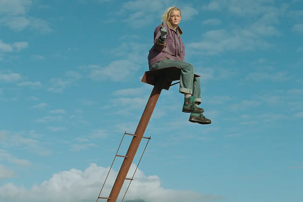 Eine Person in lila Jacke und Jeans sitzt auf einer hohen, rostigen Metallleiter vor einem blauen, wolkenverhangenen Himmel und streckt trotzig den Mittelfinger in die Kamera - eine kühne Darstellung inmitten der Echos von The Love that Remains.